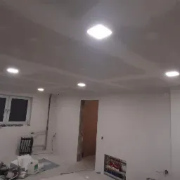 Wnętrze w trakcie tynkowania: widoczne białe ściany po nałożeniu gładzi, sufit z zamontowanymi kwadratowymi lampami LED, okno z czarną szybą, grzejnik żeberkowy i porozrzucane narzędzia malarskie...