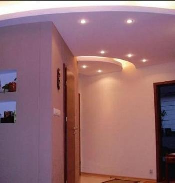 Korytarz z podwieszanym sufitem z oświetleniem punktowym, ściany w odcieniach beżu, widoczne drzwi i dekoracyjne wnęki w ścianie.