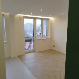 Jasne wnętrze po remoncie z parkietem ułożonym w jodełkę, widoczne niedokończone podłączenie elektryczne do oświetlenia sufitowego.