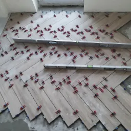 Układanie podłogi z płytek imitujących drewno w jodełkę, z użyciem poziomicy i systemu poziomowania płytek z czerwonymi klipsami i niebieskimi klinami, widoczne surowe podłoże.