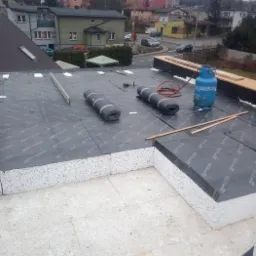 Prace dekarskie na płaskim dachu: rozłożone rolki papy termozgrzewalnej, butla gazowa i elementy drewniane na tle zabudowy miejskiej w pochmurny dzień.