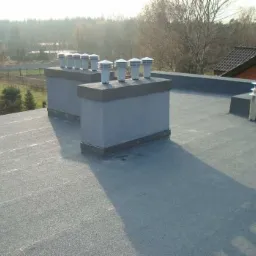 Płaski dach pokryty papą termozgrzewalną z dwoma kominami wentylacyjnymi z nasadami oraz dwoma kominami stalowymi w tle, widoczne obróbki blacharskie.