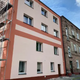 Odnowiona elewacja budynku wielorodzinnego z pomarańczowymi pasami, widoczne rusztowanie i sąsiedni budynek z cegły.