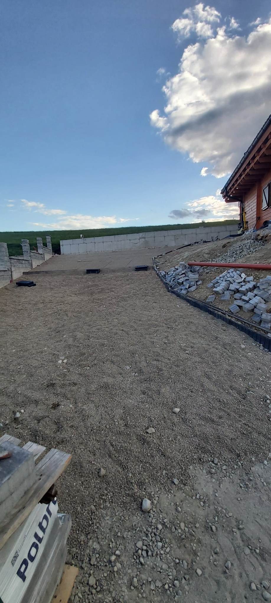Przygotowany teren pod przyszłą nawierzchnię z kostki, widoczne obrzeża, podsypka, paleta z kostką Polbruk oraz fragment drewnianego domu w tle.