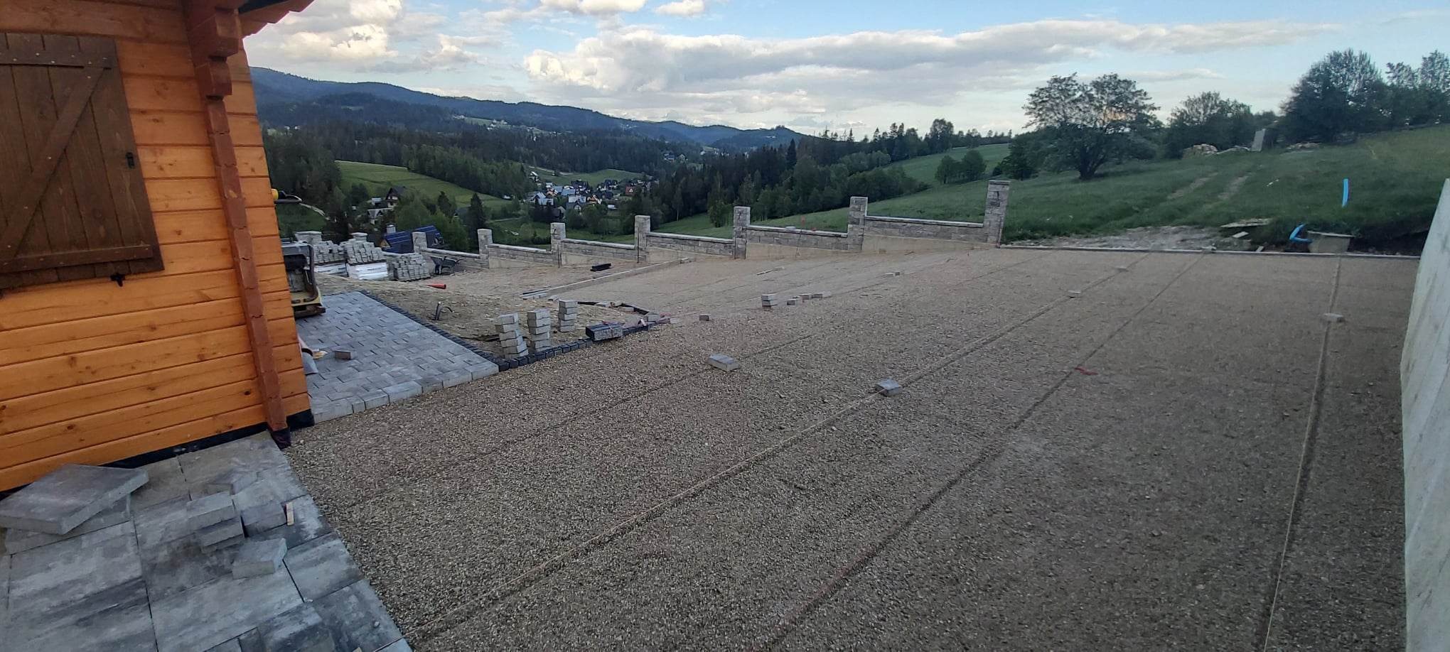 Przygotowanie podłoża pod nawierzchnię z kostki brukowej na tle malowniczego krajobrazu górskiego, widoczne ułożone obrzeża i wstępnie ułożona kostka przy drewnianym domku.