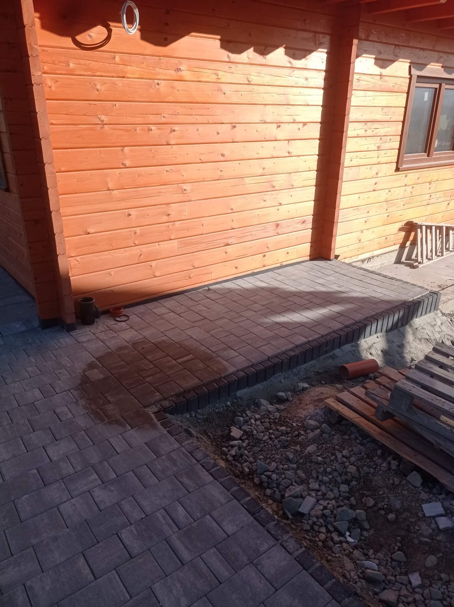 Ułożona kostka brukowa przed drewnianym domem, widoczne palety i rura, elementy układanki w trakcie montażu.