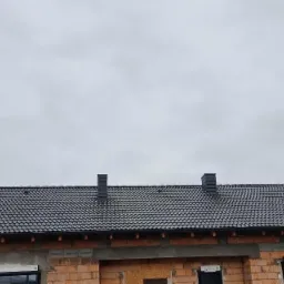 Nowy dach z czarnej dachówki ceramicznej na budynku w stanie surowym otwartym, widoczne kominy wentylacyjne.