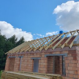 Usługi Adam Janasik - Wznoszenie więźby dachowej na murowanym domu jednorodzinnym w słoneczny dzień, widoczne przygotowane deski na dachu i na ziemi.