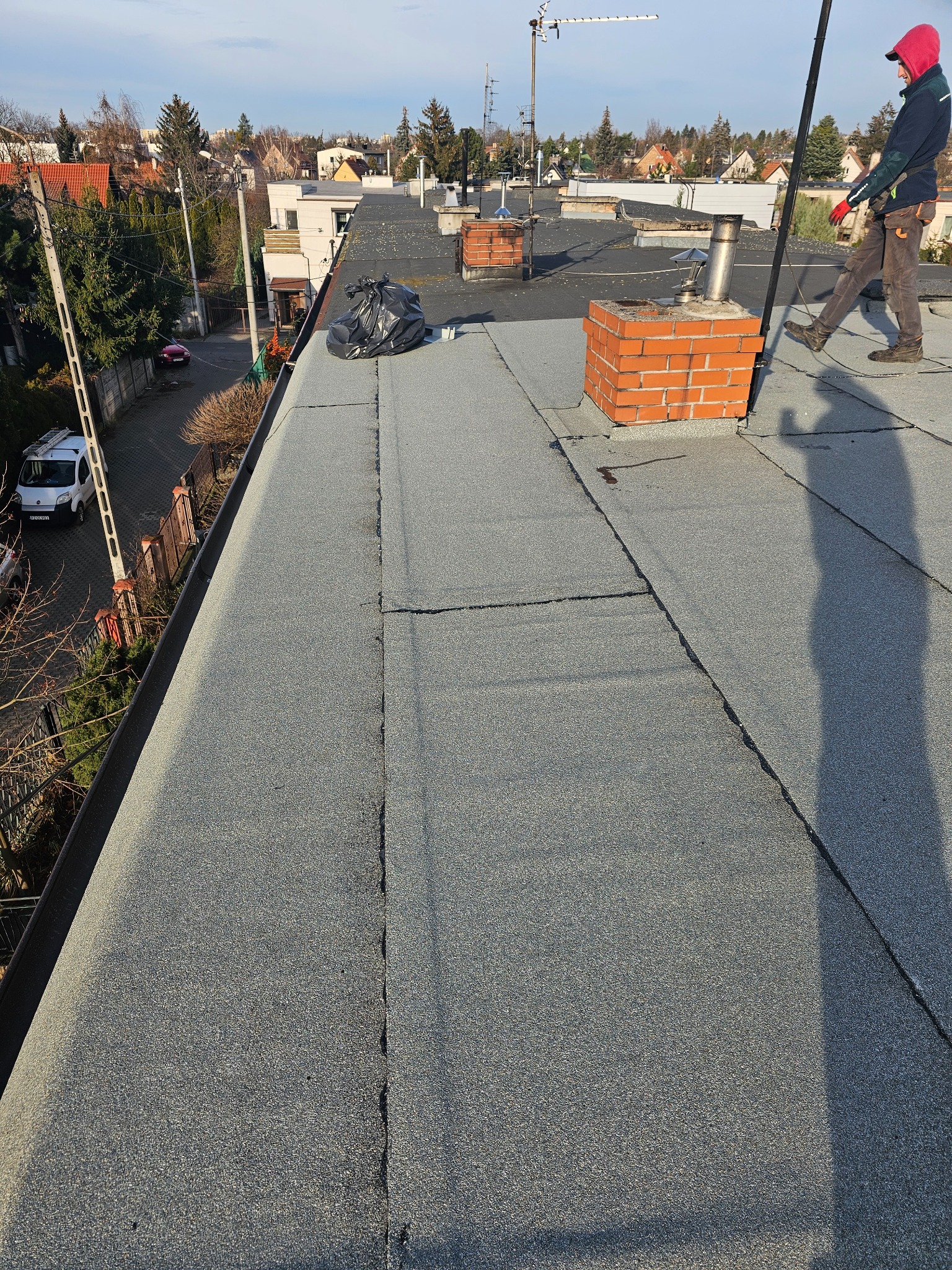 Praca na dachu pokrytym papą, widoczne pęknięcia i łączenia, komin z cegły, pracownik w odzieży roboczej i czarny worek na śmieci.