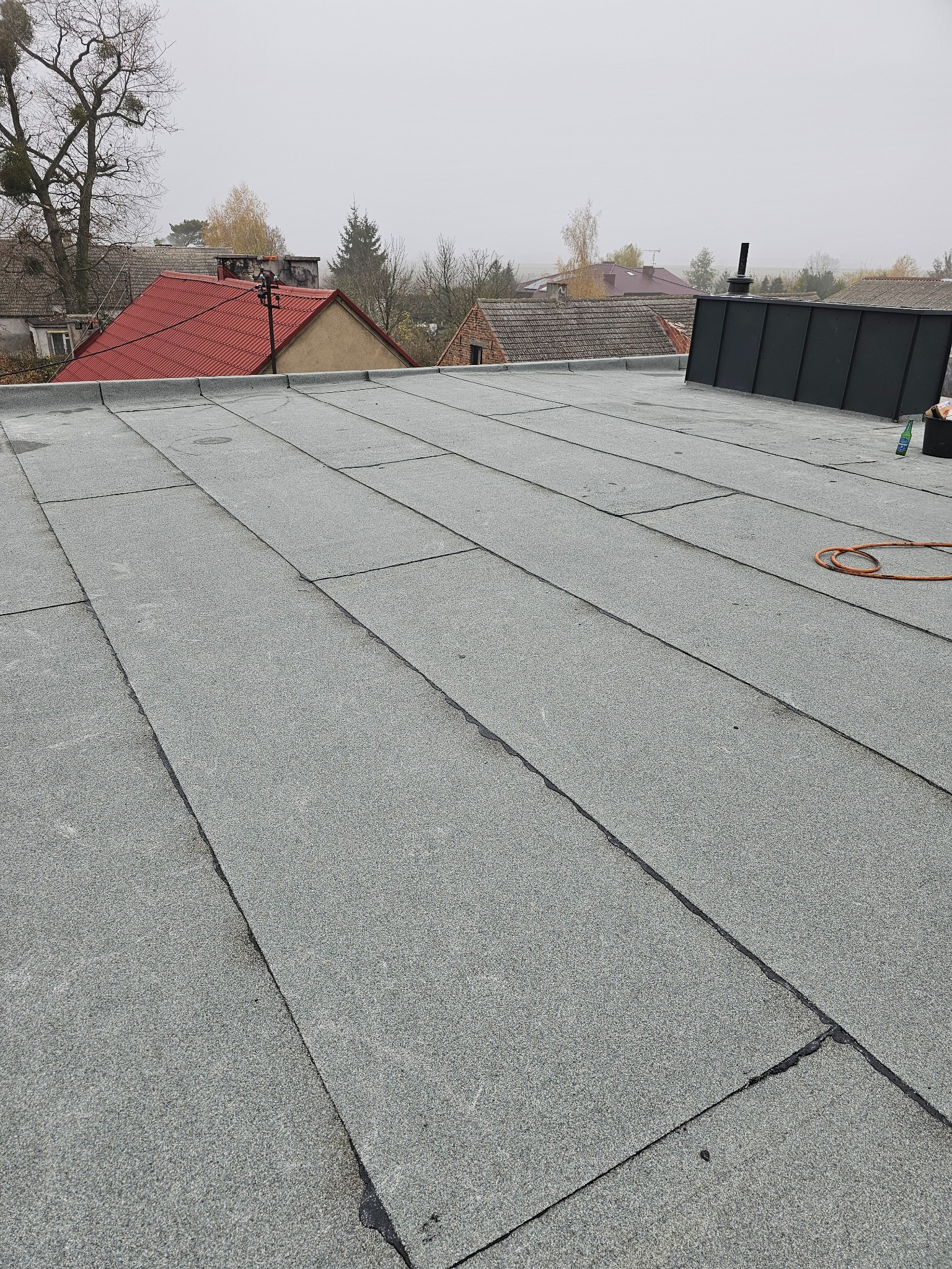 Płaski dach pokryty papą termozgrzewalną w szarym kolorze, widoczne łączenia, w tle zabudowania mieszkalne w pochmurny dzień.