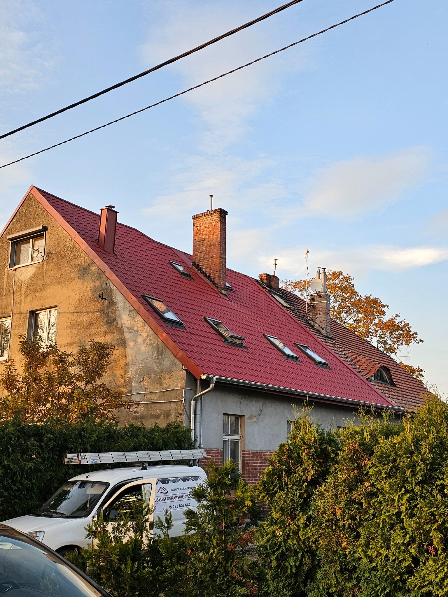 Czerwony dach z kilkoma oknami dachowymi na starszym domu z dwoma kominami, częściowo przysłonięty drzewami i z samochodem firmy dekarskiej w tle.