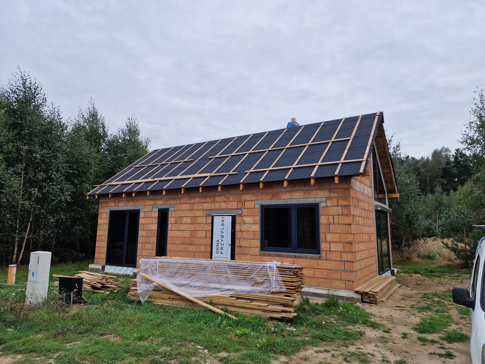 Dom w trakcie budowy z częściowo wykonanym pokryciem dachu z papy i drewnianą konstrukcją łat, widoczne materiały budowlane i otoczenie leśne.