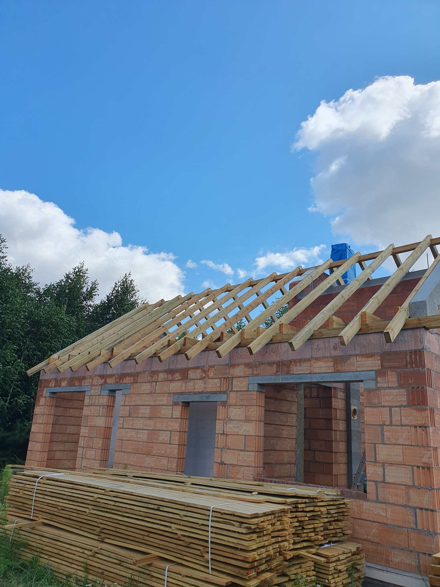 Wznoszenie więźby dachowej na murowanym domu jednorodzinnym w słoneczny dzień, widoczne przygotowane deski na dachu i na ziemi.