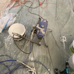 Sprzęt do natryskowego malowania ścian, w tym pistolet natryskowy, pompa malarska Classic 390, wiadro z farbą, mieszadło do farb i wiertarko-wkrętarka Makita, rozłożone na folii ochronnej.