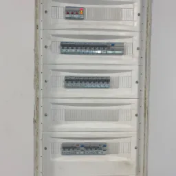 Wnętrze skrzynki rozdzielczej z bezpiecznikami, widoczne białe obudowy modułów, oznaczenia F1-F26 i Q1-Q3, oraz dioda LED sygnalizująca stan zasilania, zamontowana w ścianie z widocznymi śladami...