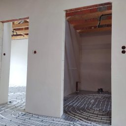 Prime Home Dewelopment sp. z o.o. sp.k. - Wnętrze w trakcie budowy z widocznymi instalacjami ogrzewania podłogowego, surowe ściany i drewniane elementy konstrukcyjne stropu.
