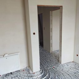 Prime Home Dewelopment sp. z o.o. sp.k. - Białe rury ogrzewania podłogowego ułożone na siatce w jasnym, surowym wnętrzu z widoczną szafką rozdzielczą przy ścianie i przejściem do kolejnego pomieszczenia.