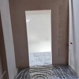 Prime Home Dewelopment sp. z o.o. sp.k. - Wnętrze w trakcie instalacji ogrzewania podłogowego, widoczne rury układane na siatce montażowej w surowym stanie, ściany otynkowane, otwór drzwiowy w głębi.