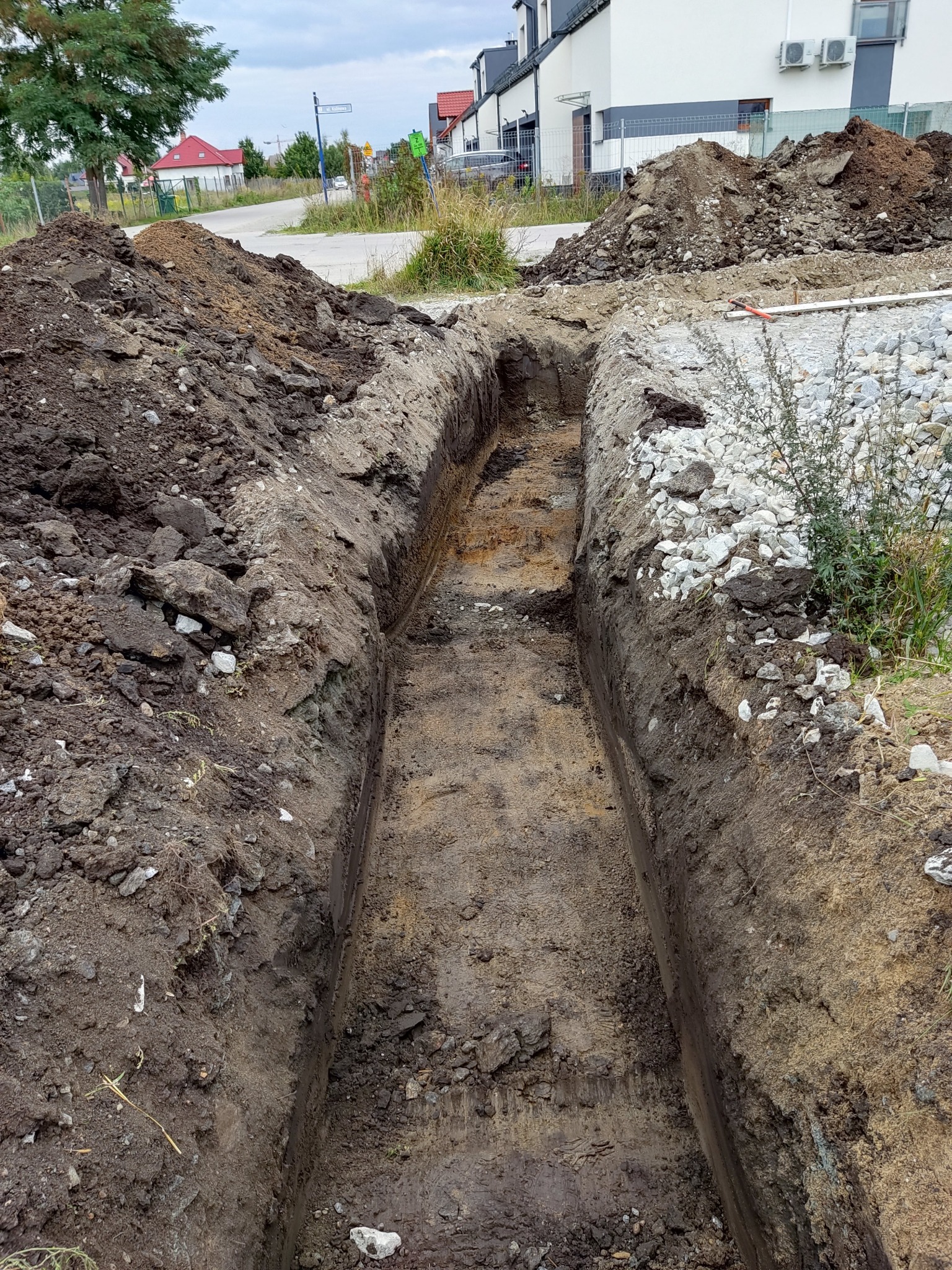 Długi, wąski wykop w ziemi, widoczne pryzmy ziemi po bokach, w tle budynki mieszkalne i fragment drogi.