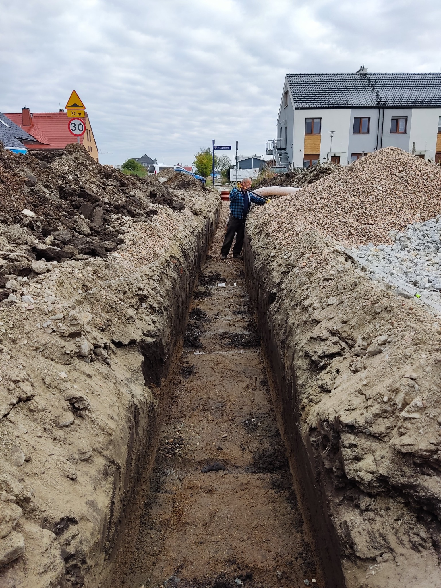 Wąski wykop w ziemi z równymi ścianami, obok hałda ziemi i żwiru, w tle budynki mieszkalne i pracownik w niebieskiej koszuli w kratę.