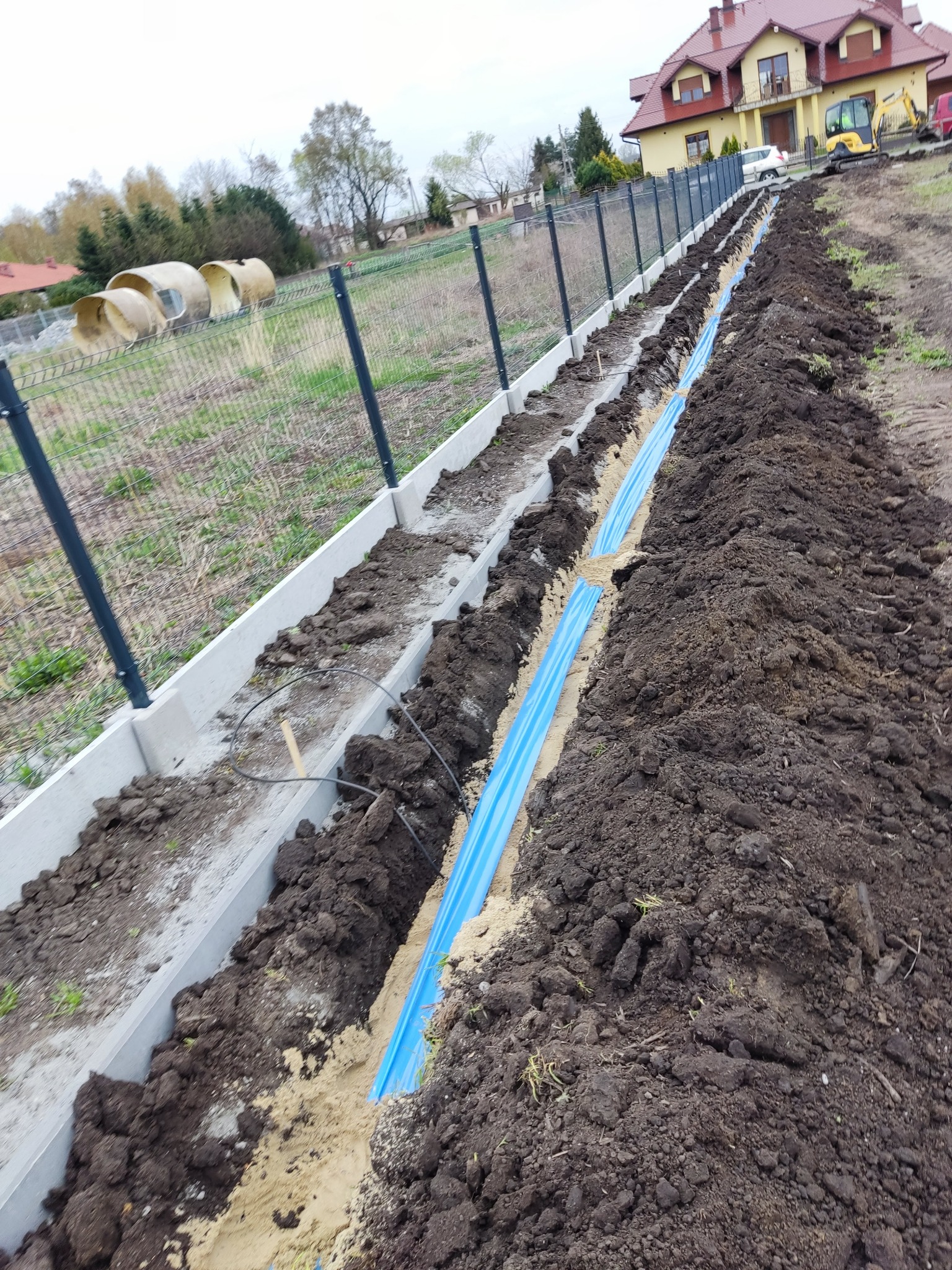 Długi wykop z ułożonym niebieskim rurociągiem, zabezpieczony betonowymi krawężnikami, w tle dom jednorodzinny i sprzęt budowlany.