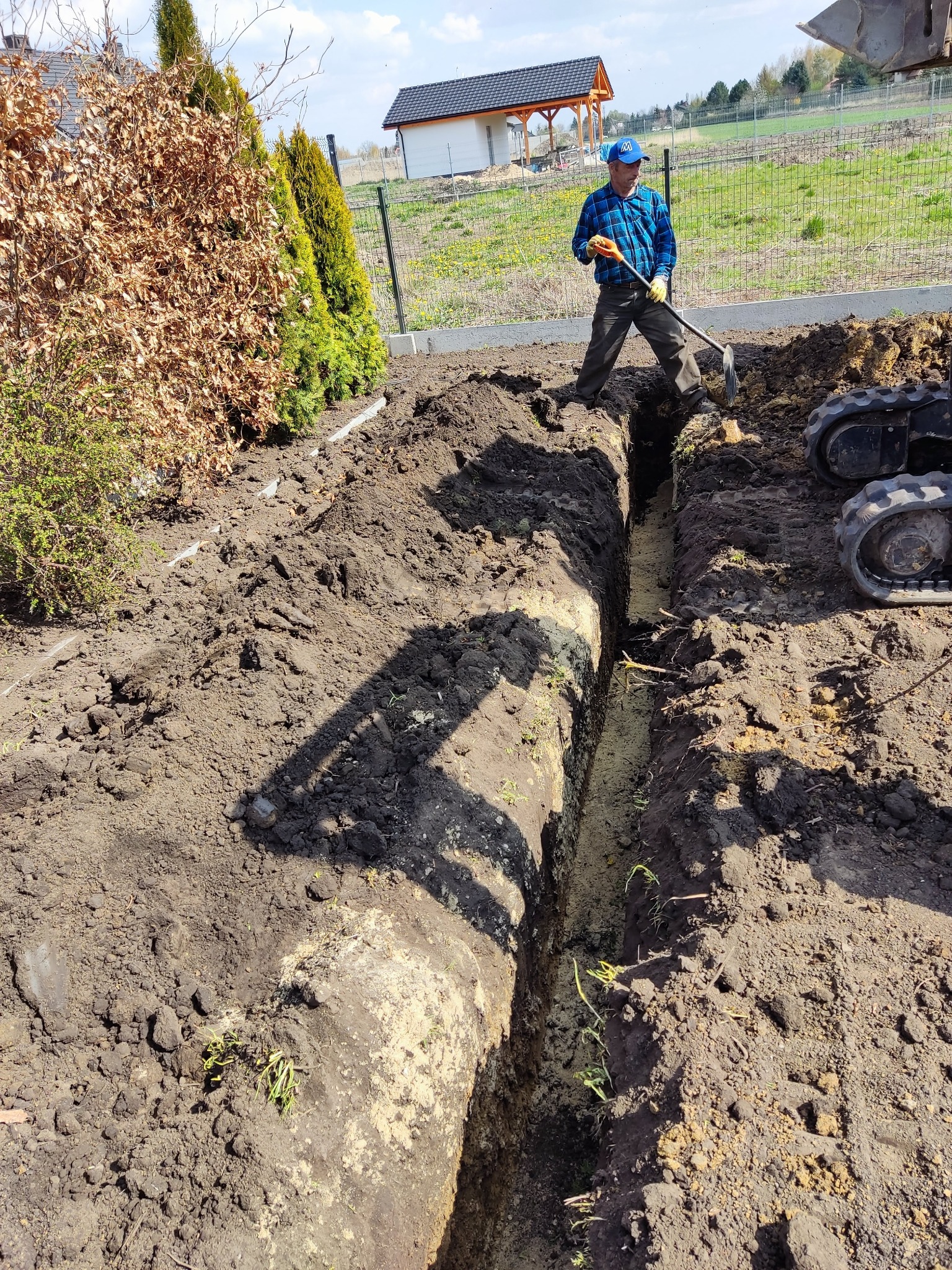 Wykopany wąski rów w ziemi, pracownik w niebieskiej koszuli z łopatą, minikoparka obok, żywopłot i dom w tle.
