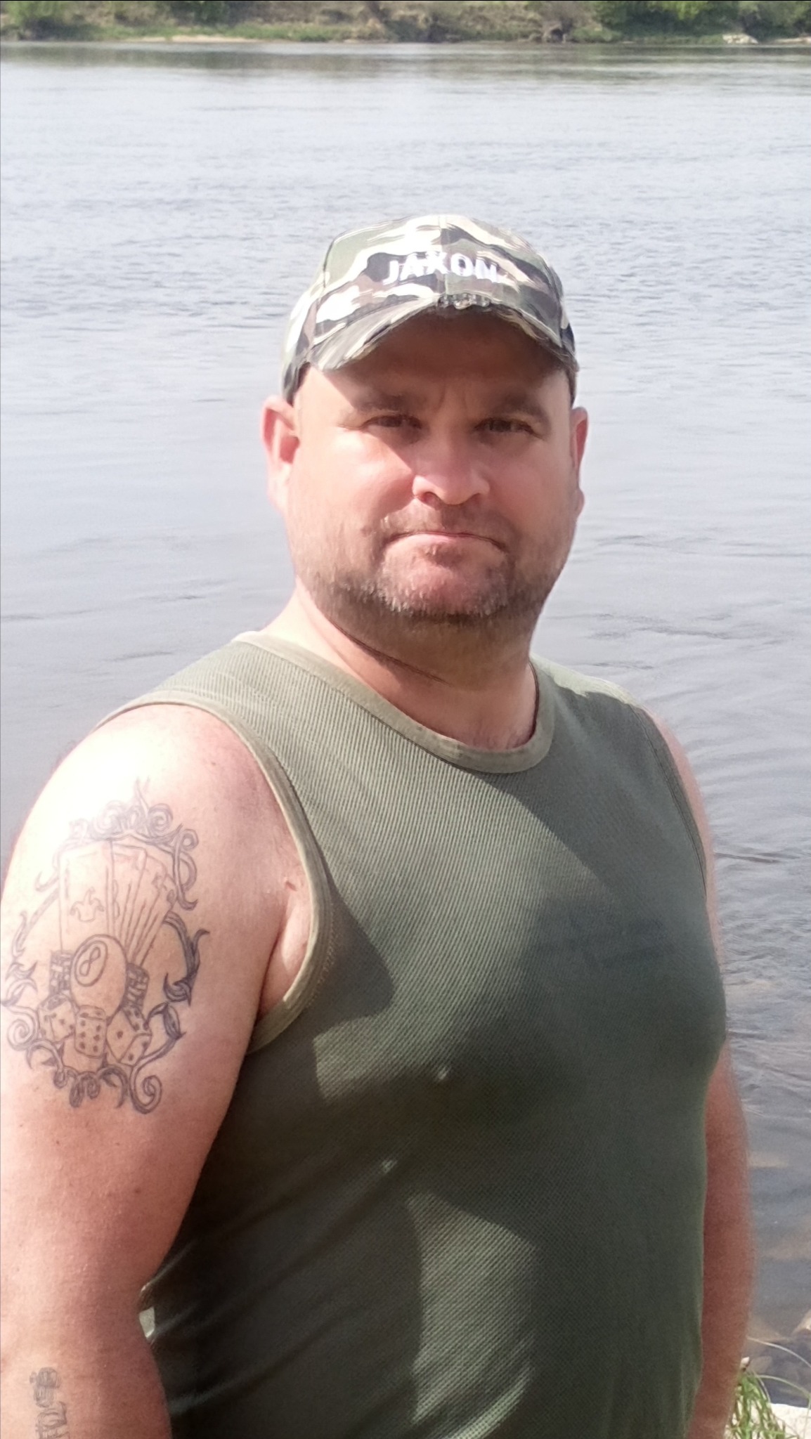 Mężczyzna w zielonym podkoszulku i czapce moro, z tatuażem na ramieniu, na tle wody.