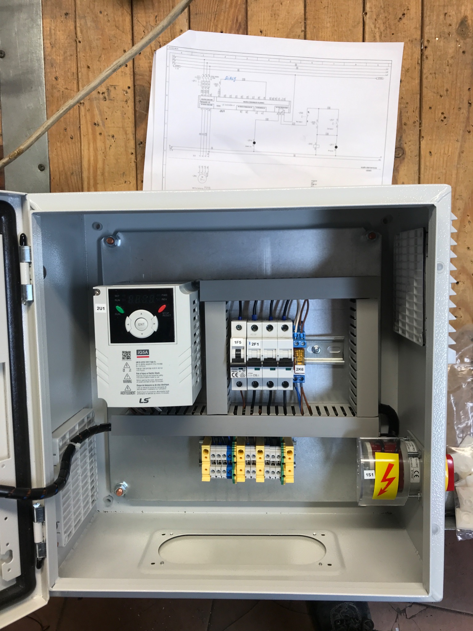 Otwarta, szara szafa sterownicza z zamontowanym falownikiem LS i bezpiecznikami, widoczne okablowanie, schemat elektryczny przywieszony obok na drewnianej ścianie.