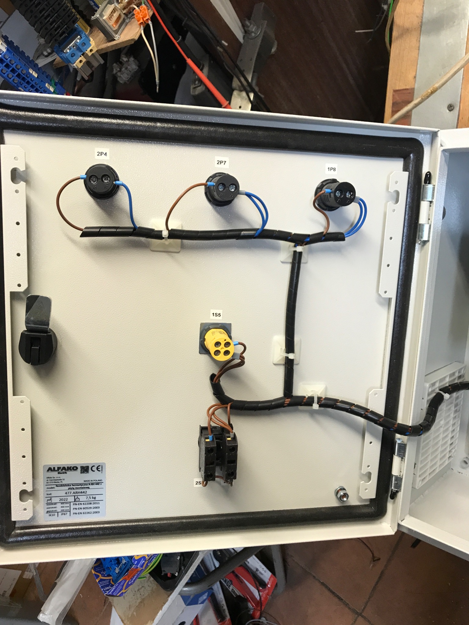 Wnętrze skrzynki elektrycznej z okablowaniem, widoczne czarne przewody w peszlu, gniazda i złączki, tabliczka znamionowa producenta Alfako.