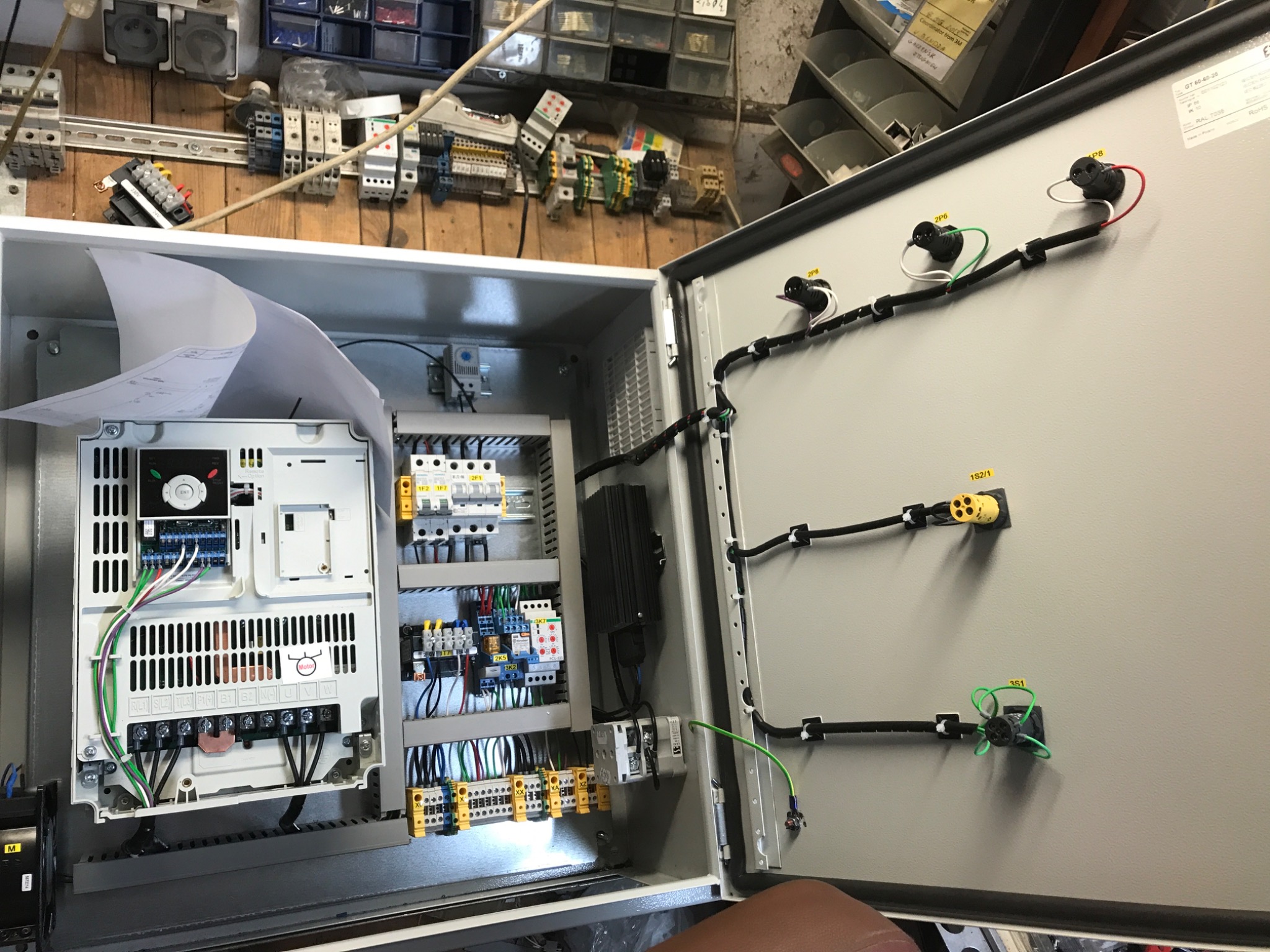 Otwarta szafa sterownicza z widocznymi elementami elektrycznymi, w tym przetwornicą częstotliwości, zabezpieczeniami, okablowaniem i schematem elektrycznym na górze. Na tylnej ścianie widoczne...