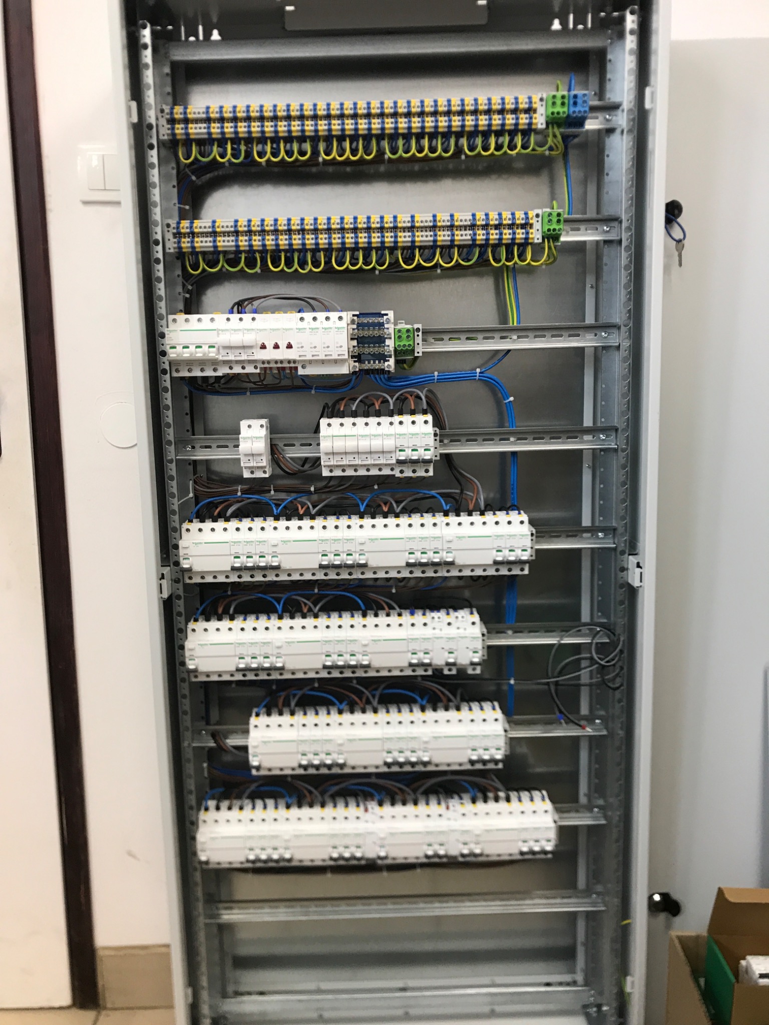 Rozdzielnica elektryczna z zamontowanymi bezpiecznikami i listwami zaciskowymi, widoczne kable w kolorach niebieskim, żółtym i zielonym, kluczyk do szafy w zamku.