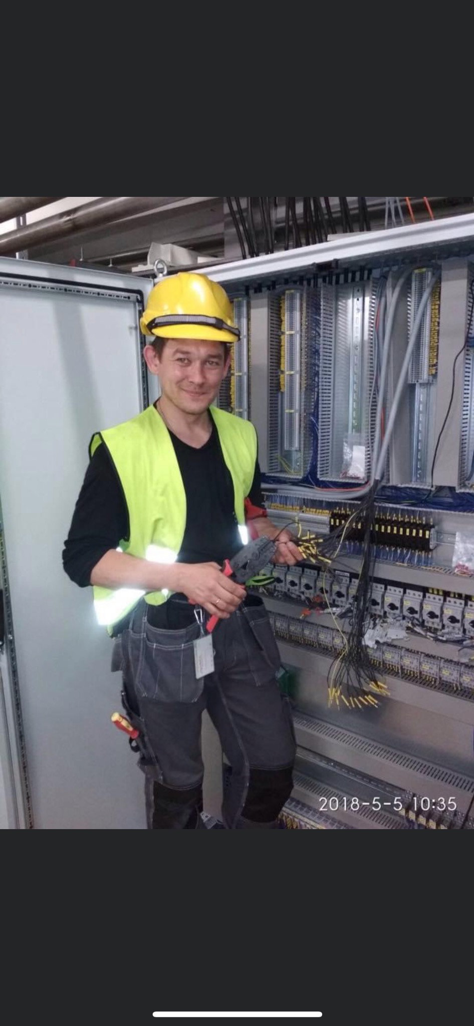 Elektryk w żółtym kasku i kamizelce, z uśmiechem trzyma w rękach obcięte przewody i kombinerki przed otwartą szafą sterowniczą z widocznymi bezpiecznikami i okablowaniem.