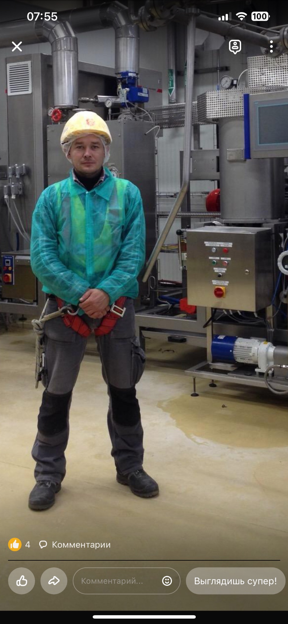 Pracownik w kombinezonie ochronnym, kasku i uprzęży, stojący na tle przemysłowej instalacji z panelami sterowania i rurami, widoczna plama cieczy na podłodze.