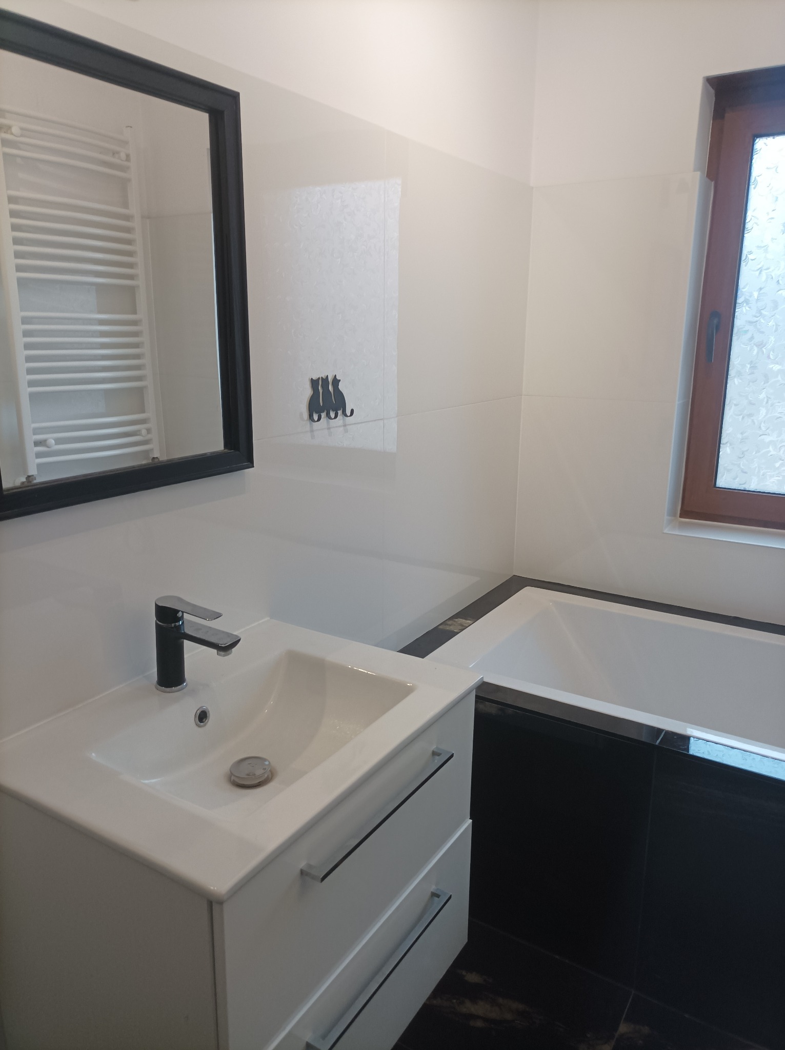 Biała umywalka z czarną baterią i szafką w nowoczesnej łazience z białymi płytkami, czarnymi akcentami i dekoracją z trzema kotami na ścianie.