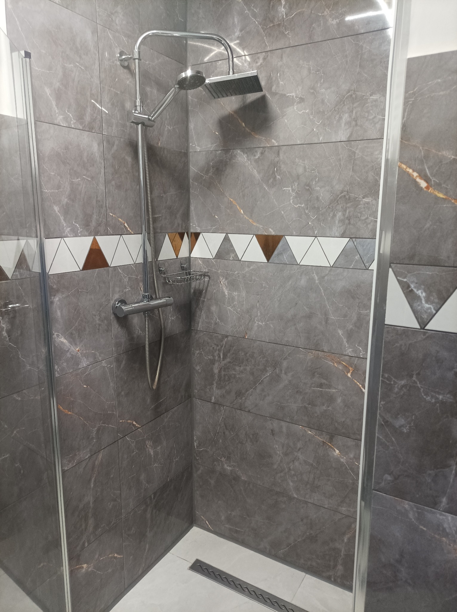 Kabina prysznicowa z szarymi płytkami imitującymi marmur, ozdobiona pasem trójkątnych dekorów w kolorach bieli, szarości i brązu, chromowana armatura i odpływ liniowy.