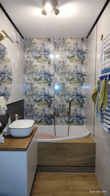 Nowoczesna łazienka po remoncie: wanna obudowana drewnem, umywalka nablatowa z baterią w kolorze złota, ściany wyłożone płytkami z motywem liści, grzejnik drabinkowy z ręcznikami.