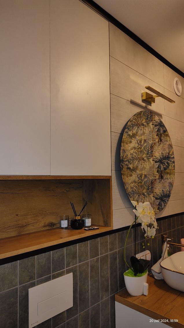 Nowoczesna łazienka z szarymi płytkami, białymi szafkami, drewnianymi elementami, okrągłym dekoracyjnym panelem na ścianie, lampą nad panelem oraz ozdobami na półce i kwiatami obok umywalki.