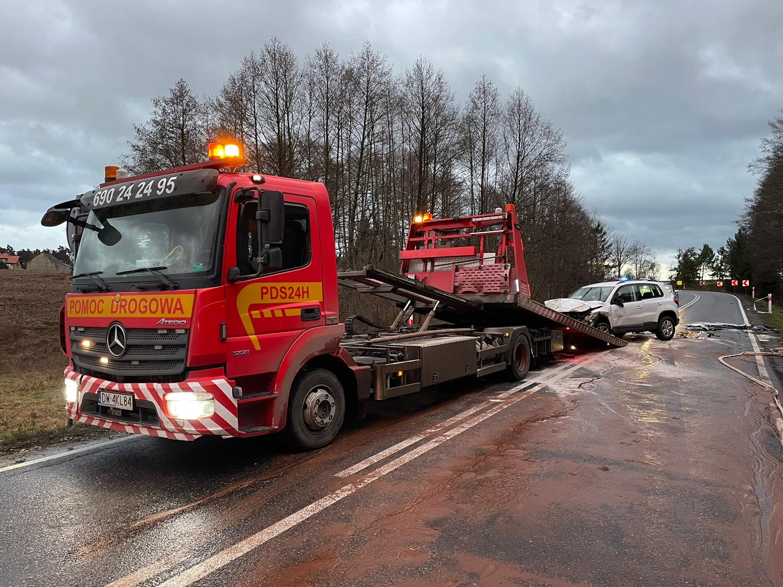Czerwona laweta Mercedes-Benz Atego z włączonymi światłami ostrzegawczymi transportuje uszkodzony, biały samochód osobowy na zakręcie drogi po wypadku, widoczne ślady płynów na asfalcie i słupki...