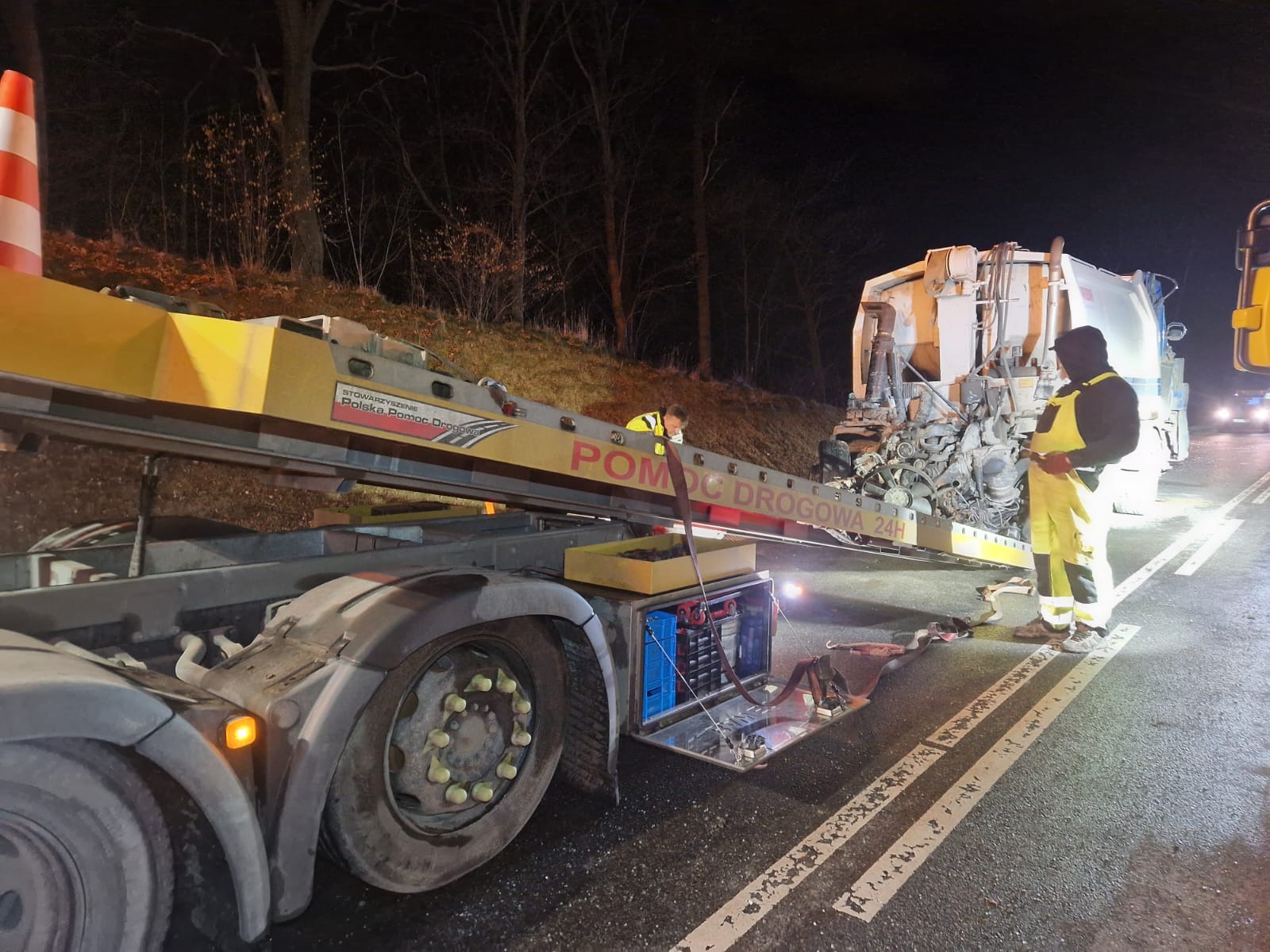Nocna akcja pomocy drogowej: uszkodzony pojazd komunalny na lawecie z napisem 'POMOC DROGOWA 24H', pracownicy w odblaskowych ubraniach, otwarte schowki z akumulatorami.