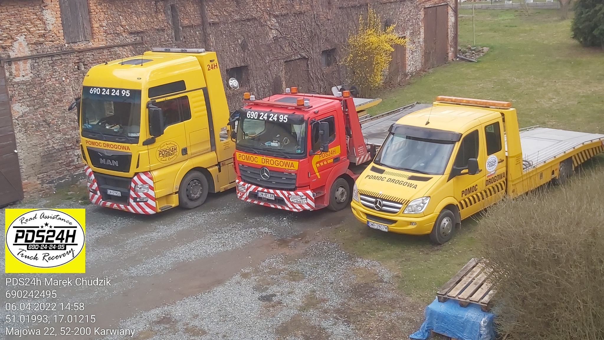 Trzy pojazdy pomocy drogowej: żółty MAN, czerwony Mercedes i żółty Mercedes Sprinter, ustawione obok siebie na żwirowym placu przed budynkiem z cegły obrośniętym bluszczem.