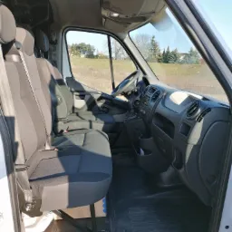 Wnętrze białego busa z widocznymi czarnymi fotelami, deską rozdzielczą i widokiem na zewnątrz na pole i drzewa w słoneczny dzień.