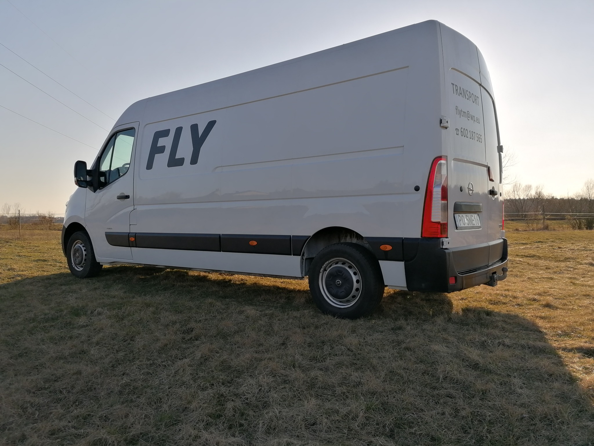 Biały bus marki Opel z napisem 'FLY' na boku, zaparkowany na polu z suchą trawą, widoczne dane kontaktowe firmy transportowej na tylnych drzwiach.