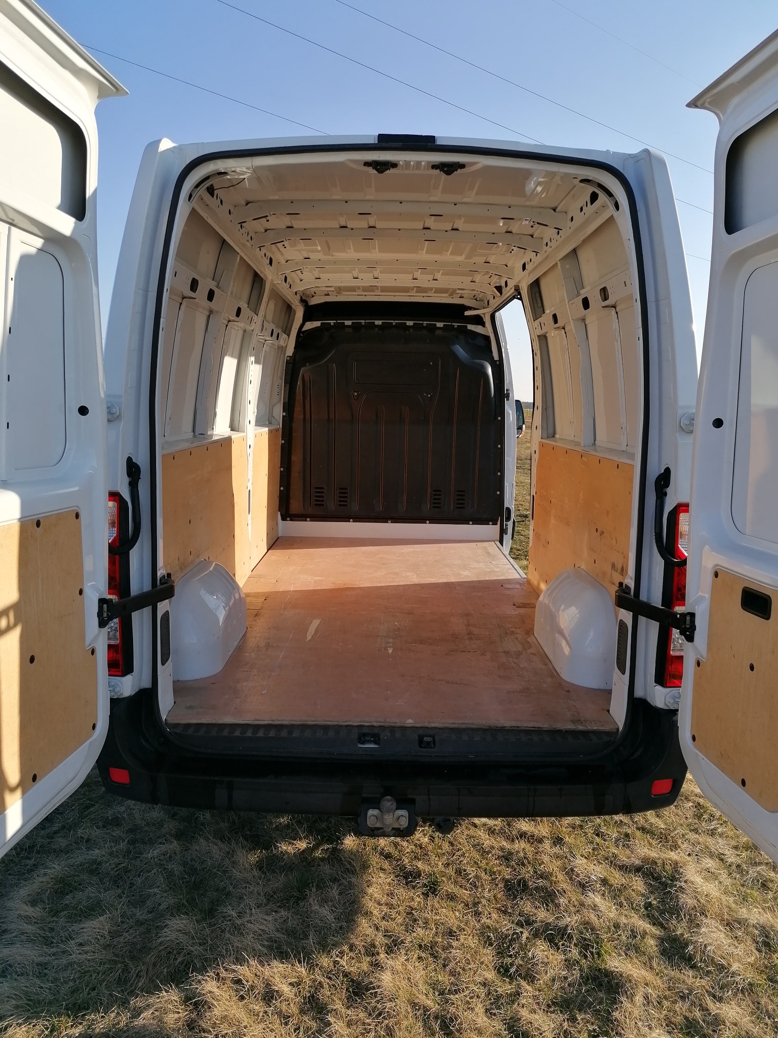 Wnętrze pustego, białego busa z otwartymi drzwiami, wyłożonego płytami z jasnego drewna, widok z tyłu na tle suchej trawy i błękitnego nieba.