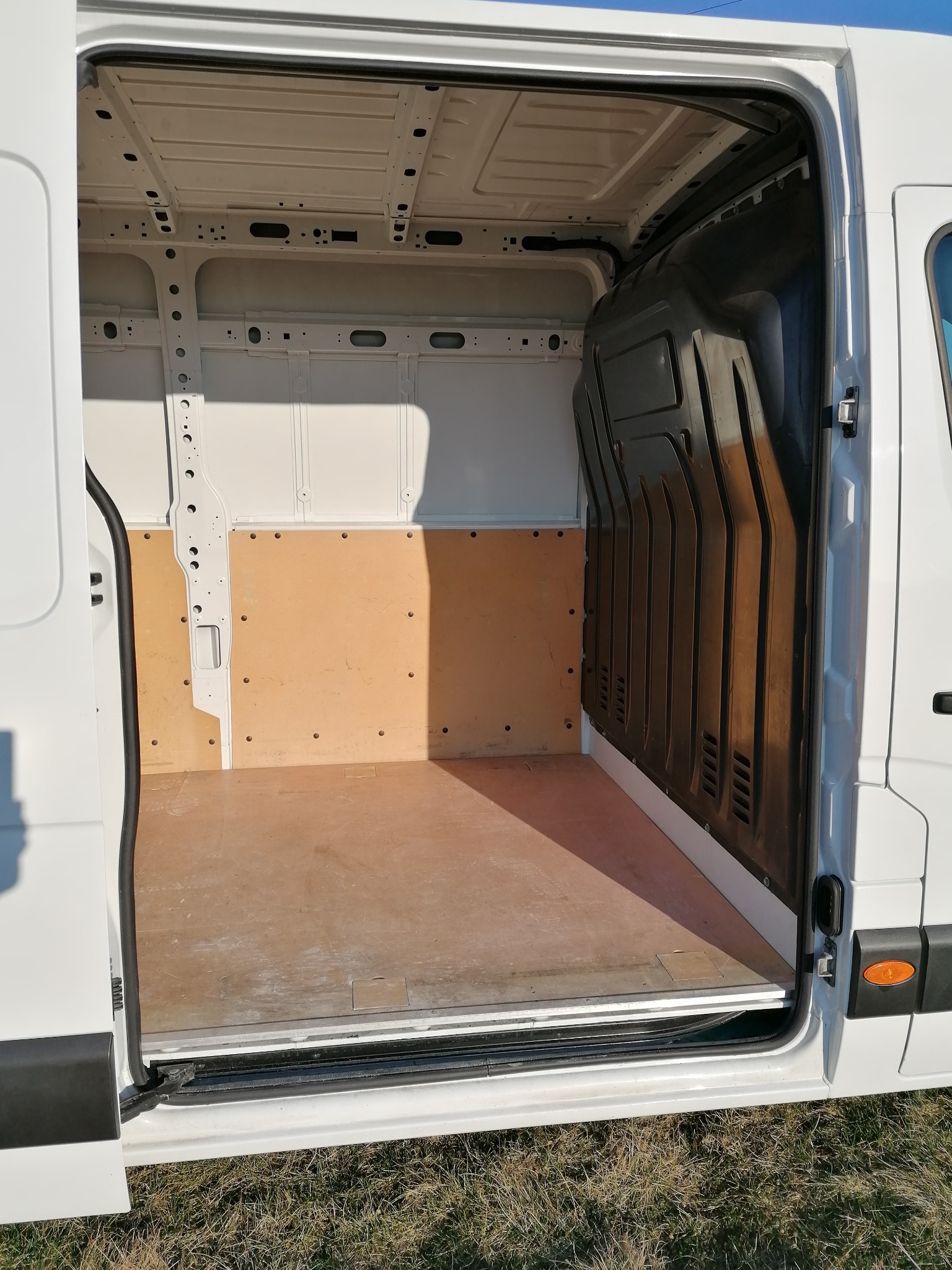 Wnętrze pustego, białego busa z otwartymi drzwiami bocznymi, widoczna drewniana podłoga i panele ochronne na ścianach.