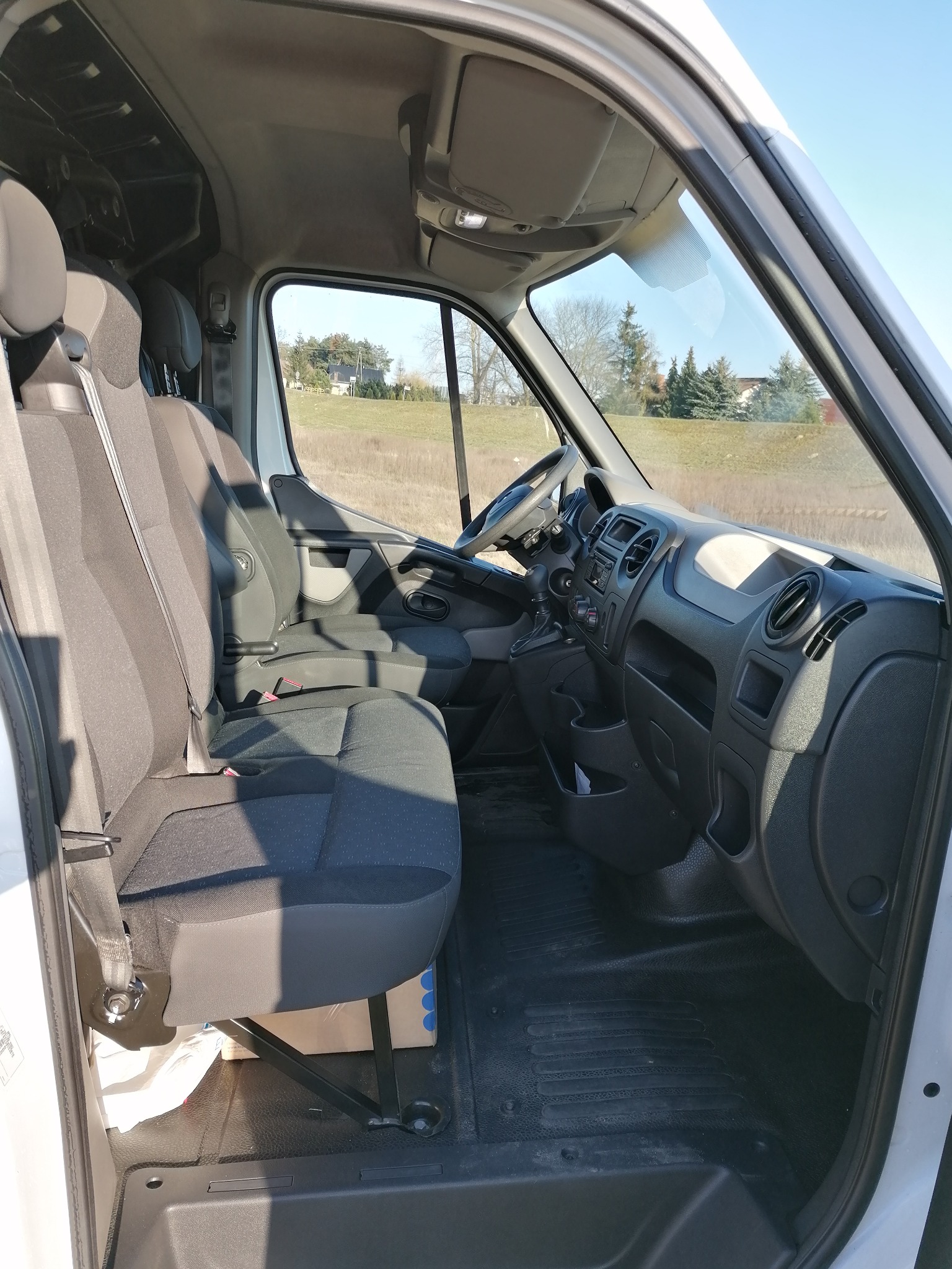 Wnętrze białego busa z widocznymi czarnymi fotelami, deską rozdzielczą i widokiem na zewnątrz na pole i drzewa w słoneczny dzień.