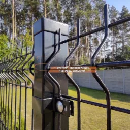 Czarny, metalowy płot panelowy z prostokątnym słupkiem, widoczne mocowanie panela, w tle drzewa i betonowe ogrodzenie.