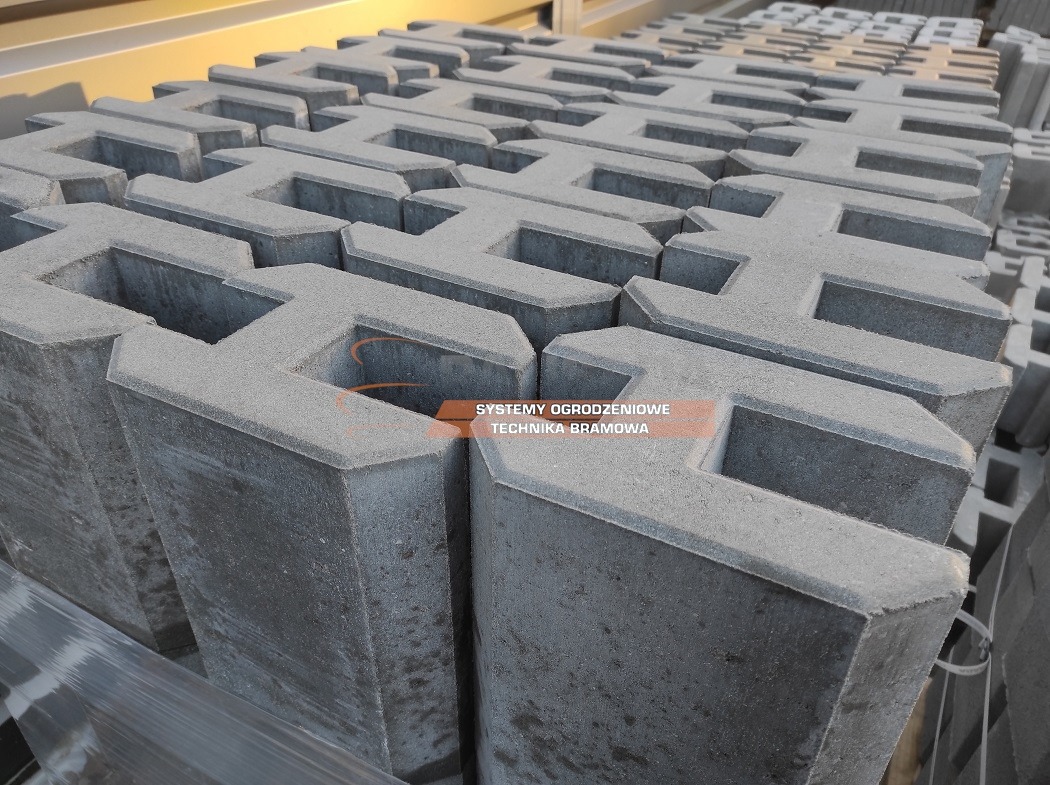Szare, betonowe elementy ogrodzeniowe ułożone jeden na drugim, tworzące wzór przypominający labirynt, widoczne w magazynie lub składzie budowlanym, opakowane w folię.