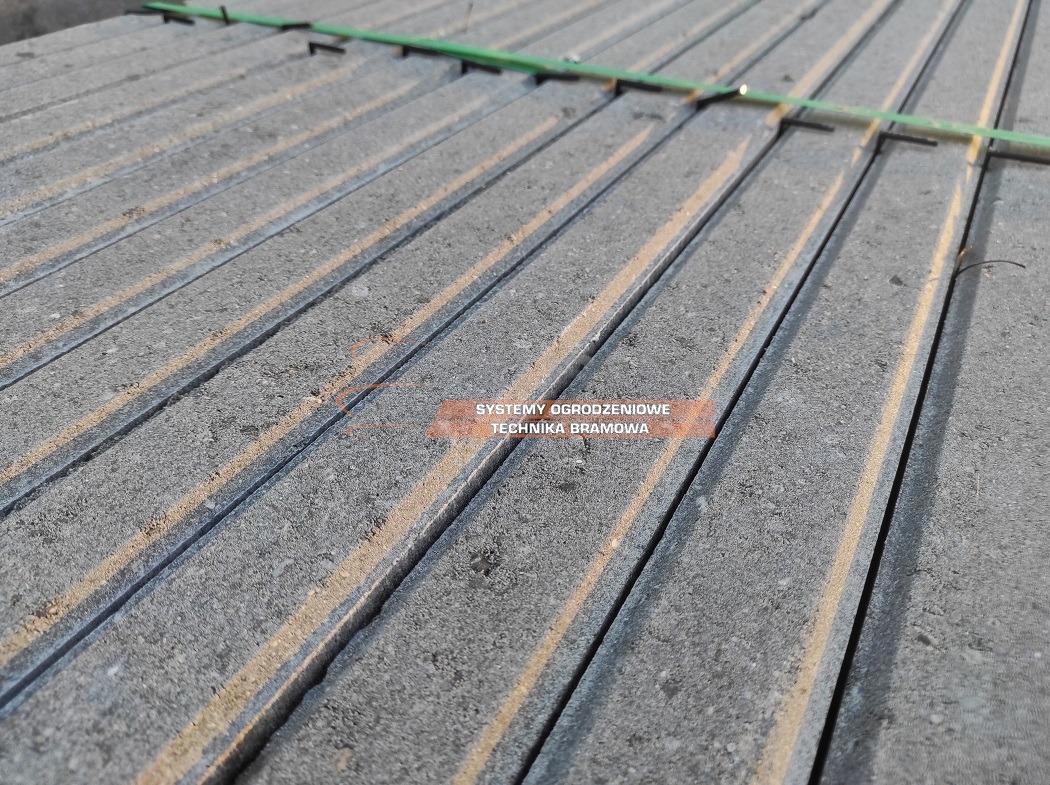 Stos szarych betonowych elementów ogrodzeniowych, ułożonych równolegle i oddzielonych ciemnymi, wąskimi szczelinami, z zielonymi plastikowymi przekładkami na górze.