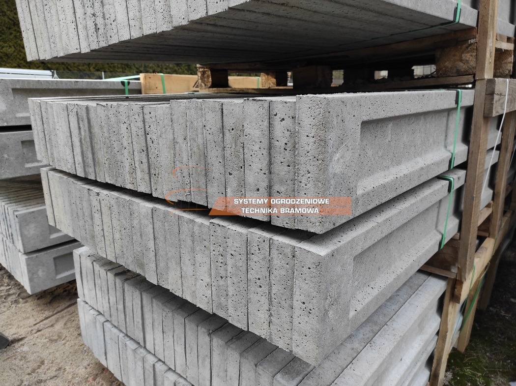 Szare, betonowe elementy ogrodzeniowe ułożone warstwami na palecie, widoczne pory i faktura betonu.