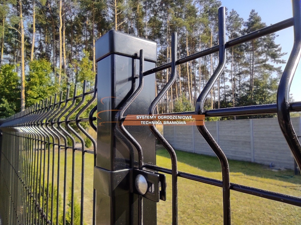 Czarny, metalowy płot panelowy z prostokątnym słupkiem, widoczne mocowanie panela, w tle drzewa i betonowe ogrodzenie.